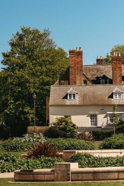 The image shows the upper section of a traditional brick house with a garden in the foreground. The house features a red brick exterior with a white-framed sash window below, and the roofline is accen